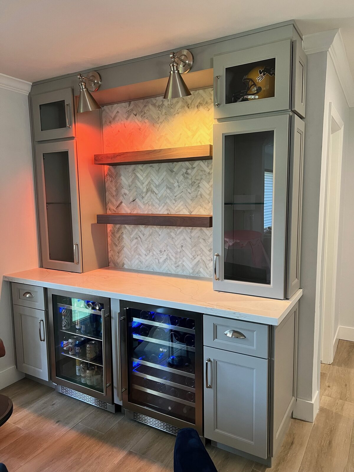 Built-in bar with gray cabinets, glass doors, floating walnut shelves, herringbone marble backsplash and two beverage coolers